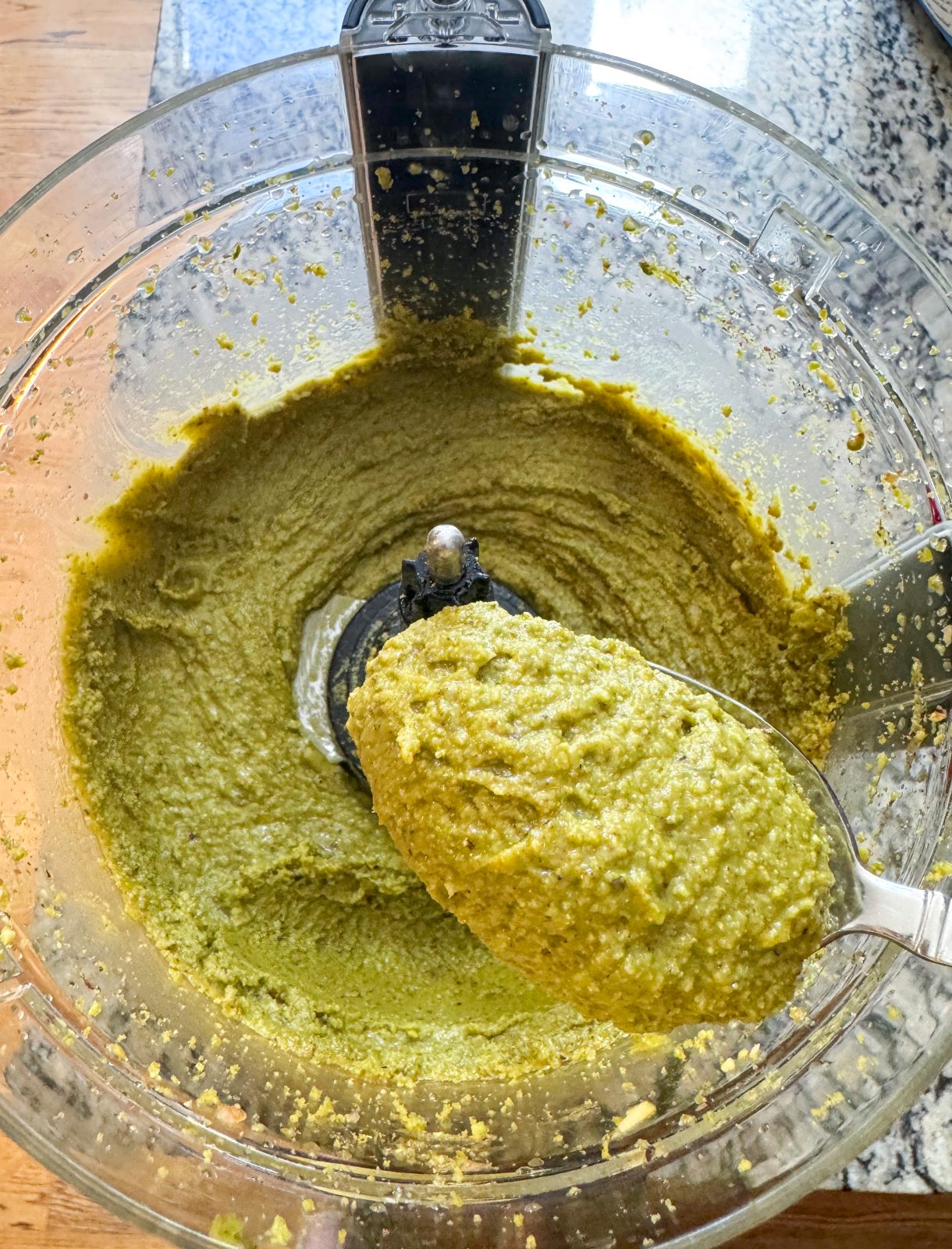 Recipe photo showing texture of pistachio butter in the bowl of the food processor. spoonful being held up to show texture.