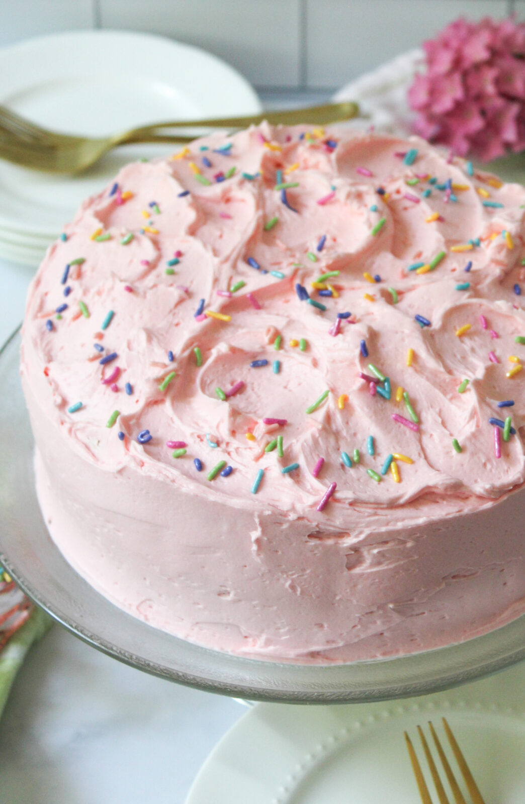 Pastel pink frosted cake with festive colorful sprinkles on top. Smooth sides of cake with fluffy swirls on top. Homemade style. Cake is on a glass pedestal with a stack of plates, forks and the bloom of a pink hydrangea in the background.