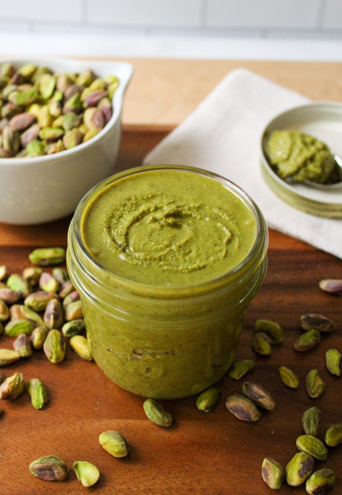 Recipe Photo: Freshly ground pistachio butter in a 8 ounce mason jar. Pretty green color. Jar is on a wooden tray with a dish of spilled pistachios and a spoonful of butter on a white dish in the background.