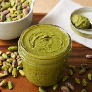Recipe Photo: Freshly ground pistachio butter in a 8 ounce mason jar. Pretty green color. Jar is on a wooden tray with a dish of spilled pistachios and a spoonful of butter on a white dish in the background.