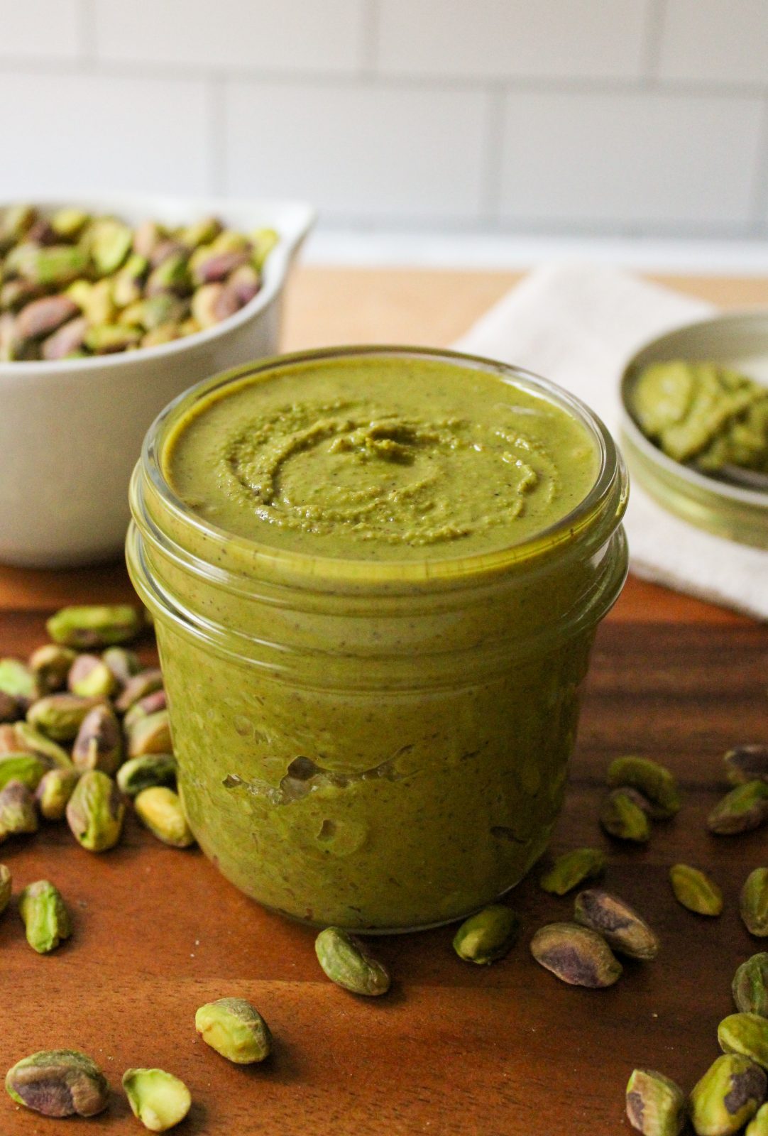 Recipe Photo: Freshly ground pistachio butter in a 8 ounce mason jar. Pretty green color. Jar is on a wooden tray with a dish of spilled pistachios and a spoonful of butter on a white dish in the background.