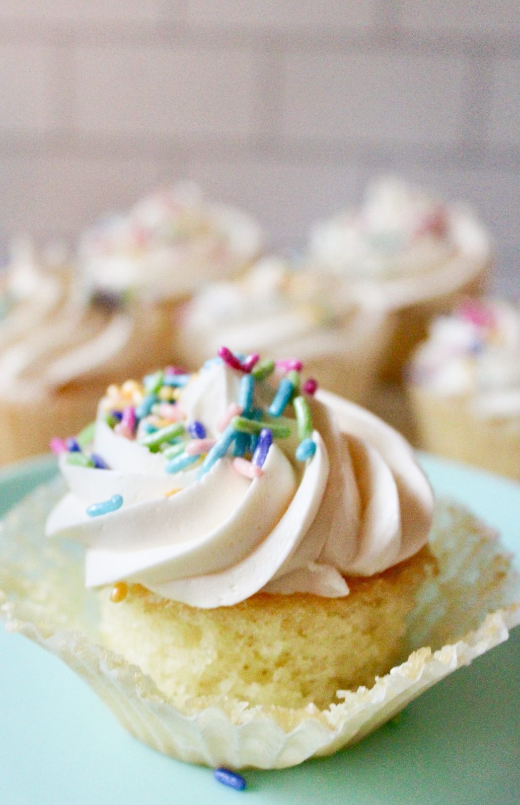 Recipe Photo for Less Sweet American Buttercream Frosting. Vanilla cupcake with piped swirl of vanilla buttercream topped with pastel sprinkles.  Cupcake liner is pulled back to reveal moist cake.  More cupcakes in back ground.