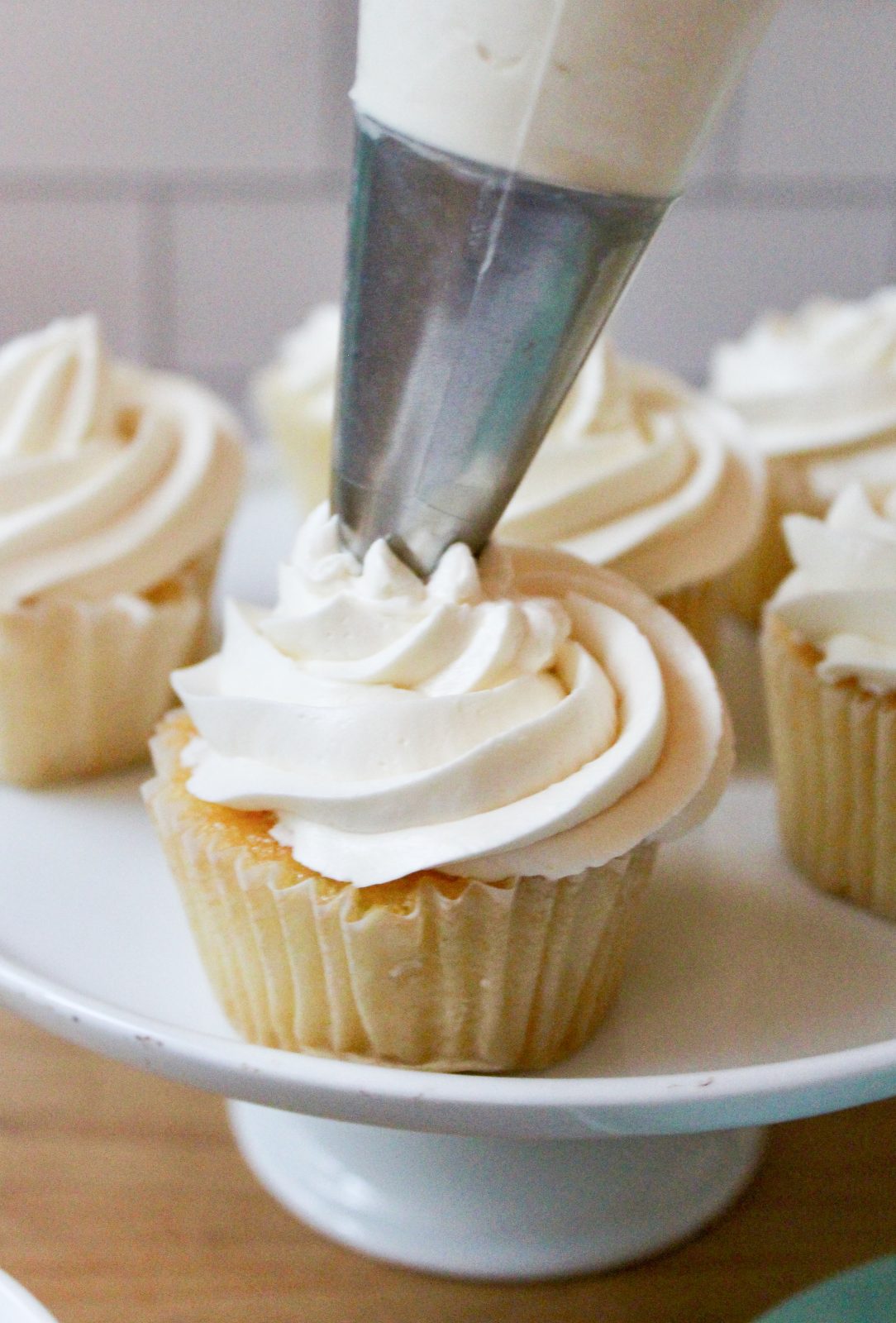 Recipe Photo for Less Sweet American Buttercream Frosting. Piping bag with metal star tip piping white buttercream onto a vanilla cupcake.  More cupcakes on white pedestal in the background.