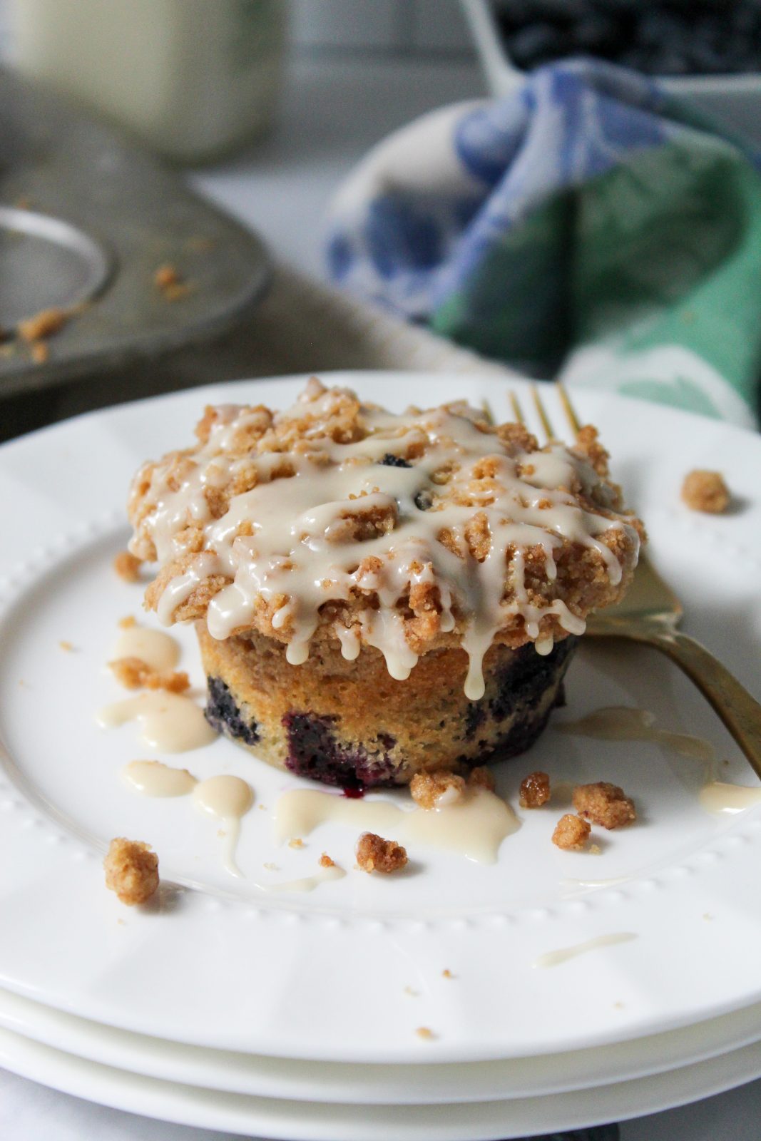 Recipe Photo - Blueberry Muffins with Cinnamon Streusel and Maple Glaze. Portrait orientation, close up showing a blueberry muffin with cinnamon streusel and dripping maple glaze on a white scalloped plate with a gold fork. In the background a cloth blue and green napkin and muffin tin.