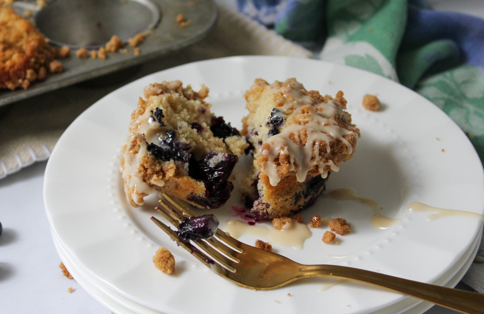Recipe Photo - Blueberry Muffins with Cinnamon Streusel and Maple Glaze. Landscape orientation showing a blueberry muffin split in half from the top on a white scalloped plate. In the background a cloth blue and green napkin and muffin tin.