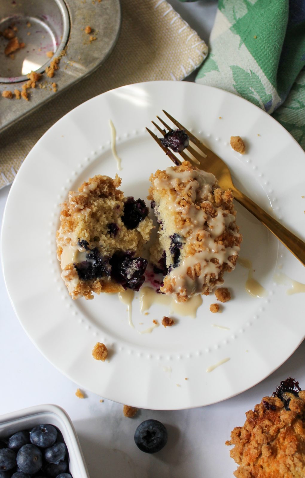 Recipe Photo - Blueberry Muffins with Cinnamon Streusel and Maple Glaze. Portrait orientation showing a blueberry muffin split in half from the top on a white scalloped plate with a gold fork. In the background a cloth blue and green napkin and muffin tin, a white ceramic basket of blueberries and another baked muffin.