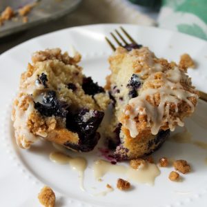 Recipe Photo - Blueberry Muffins with Cinnamon Streusel and Maple Glaze. Portrait orientation, close up showing a blueberry muffin split in half with cinnamon streusel and dripping maple glaze on a white scalloped plate with a gold fork. In the background a cloth blue and green napkin and muffin tin.