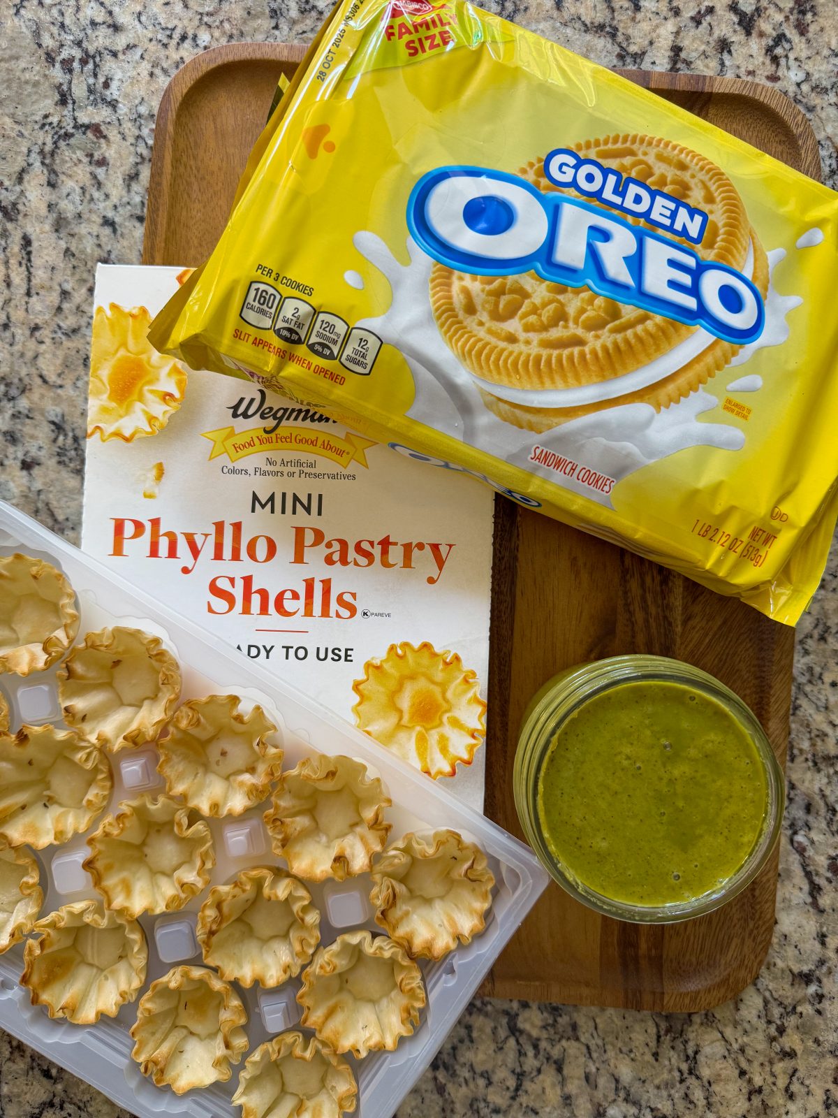 Recipe photo for Dubai Style Pistachio Oreo Truffles, Shows the ingredients laid out on a countertop. Golden Oreo package, Phyllo Pastry Shells Box and plastic tray of the shells a small mason jar filled with green pistachio butter.