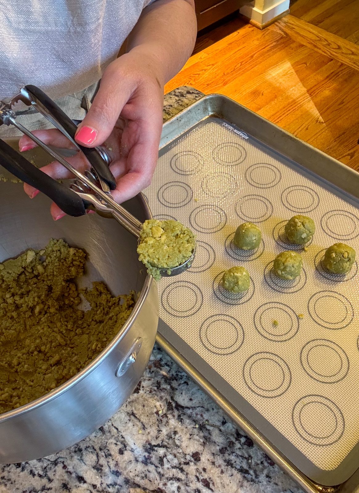 Recipe Process photo shows process of making Dubai Style Pistachio Oreo Truffles. To the side is a KitchenAid mixer bowl holding the crumbly green mixture. A sheet tray with a silicone mat with some rolled balls. A hand with a cookie scoop filled with the green mixture. The mixture and balls are pistachio green in color.