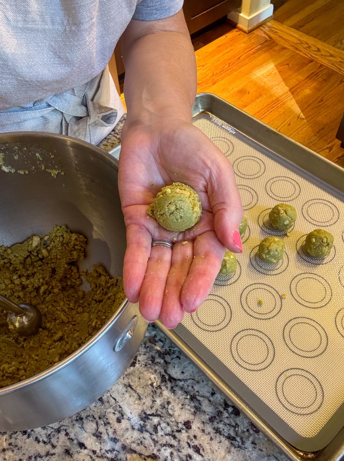 Recipe photo shows process or making Dubai Style Pistachio Oreo Truffles. To the left side is a KitchenAid mixer bowl holding the crumbly green mixture. A sheet tray with a silicone mat with some rolled balls. A hand with a freshly rolled ball. The mixture and balls are pistachio green in color.