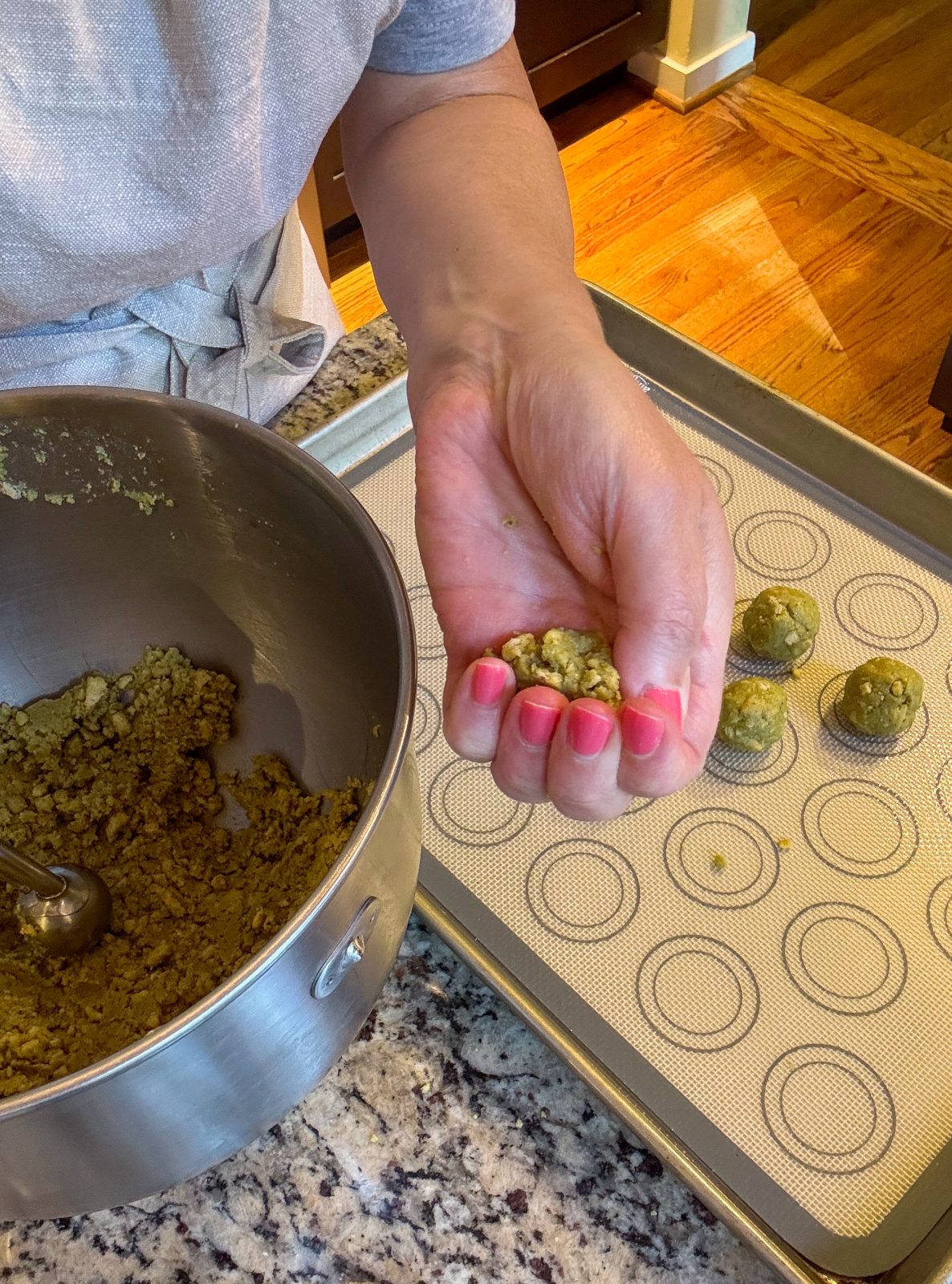 Recipe photo shows process or making Dubai Style Pistachio Oreo Truffles. To the side is a KitchenAid mixer bowl. a hand is squeezing a ball of the crumbly green mixture to get it to stick together prior to rolling the ball. A sheet tray with a silicone mat with some rolled balls. The mixture and balls are pistachio green in color.