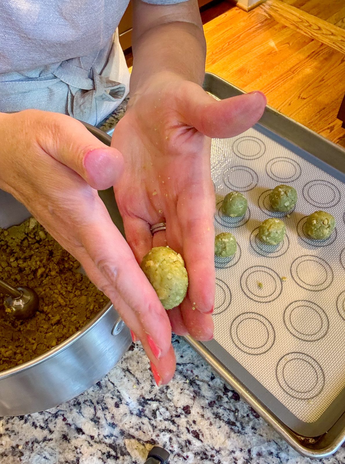 Recipe photo shows process or making Dubai Style Pistachio Oreo Truffles. To the side is a KitchenAid mixer bowl. Two hands are rolling a ball of the crumbly green mixture. A sheet tray with a silicone mat with some rolled balls. The mixture and balls are pistachio green in color.