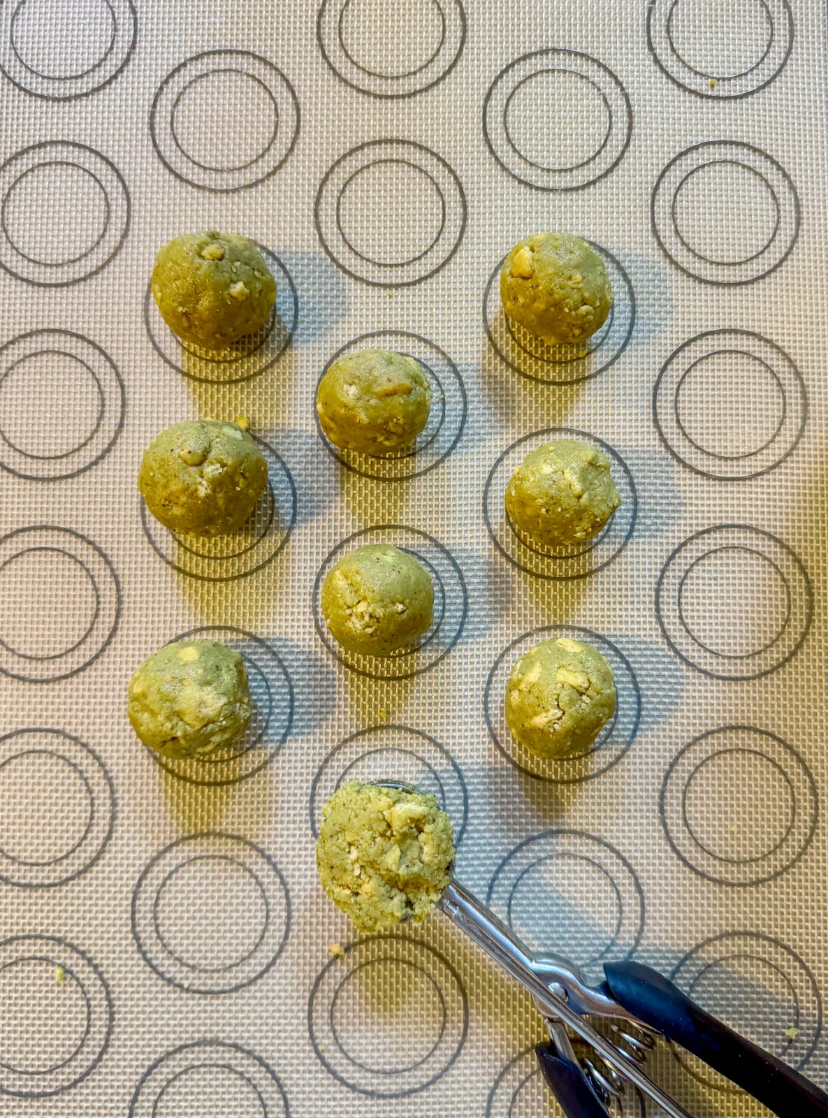 Recipe Process Photo for Dubai Style Pistachio Oreo Truffles shows silicone mat with 9 green rolled pistachio truffles and a cookie scoop.