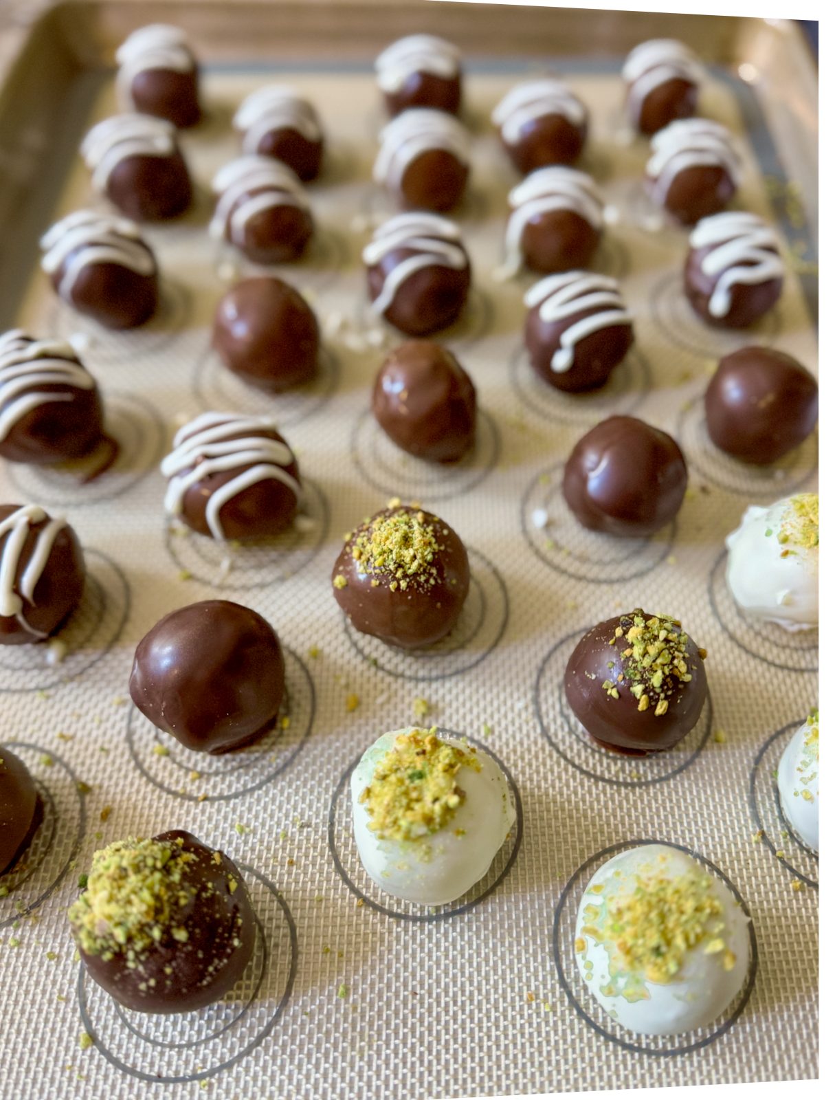 Recipe Process Video for Dubai Style Pistachio Oreo Truffles. Shows sheet tray with silicone mat filled with white, dark and milk chocolate coated pistachio truffles decorated with white chocolate drizzles or pistachio dust.