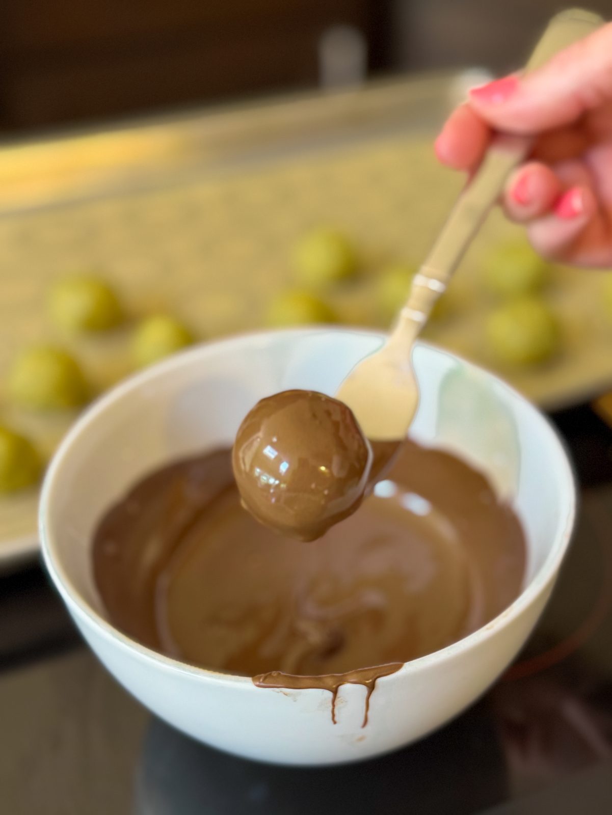 Recipe Process photo shows dipping Dubai Style Oreo Truffle Ball in a white bowl filled with melted milk chocolate. A gold fork is lifting the coated ball out of the bowl. In the background is a sheet tray filled with green balls waiting to be dipped.