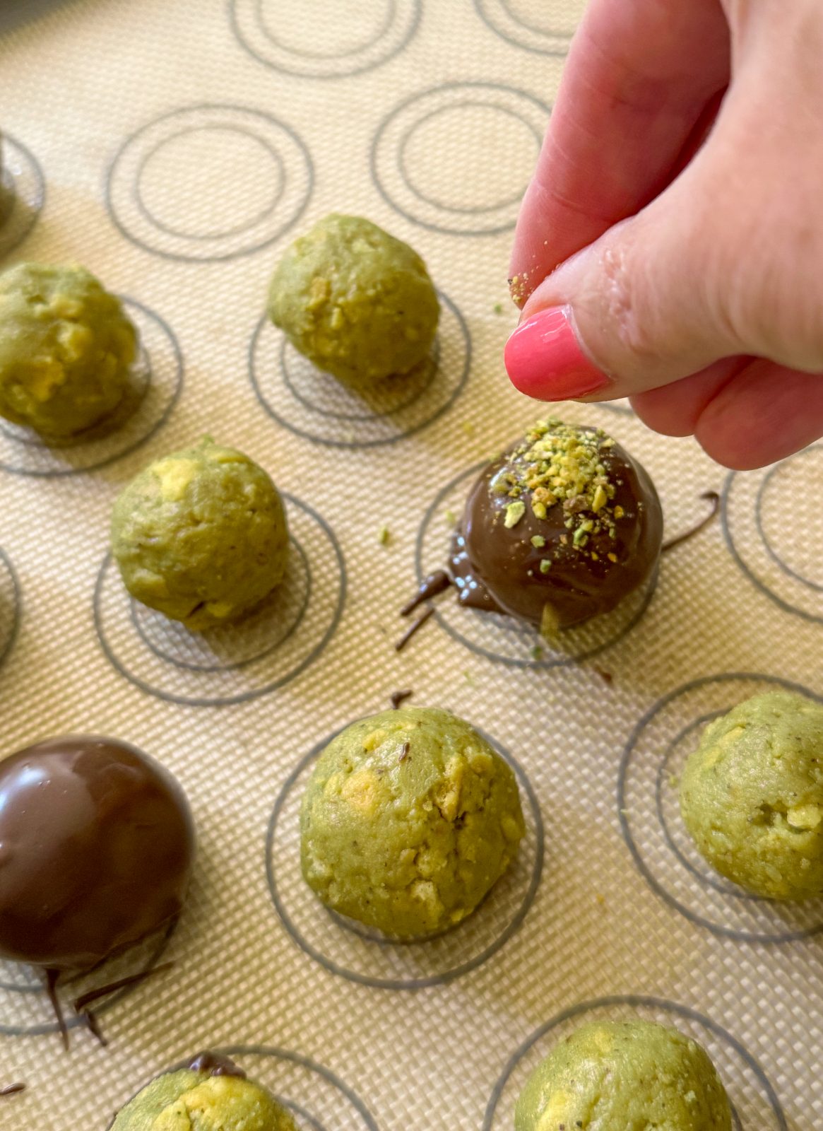 Recipe Process Photo for Dubai Style Pistachio Oreo Truffles. A silicone mat with some chocolate coated truffles and some green uncoated truffles. A hand with pink finger nails is sprinkling pistachio dust on one of the chocolate coated truffles.
