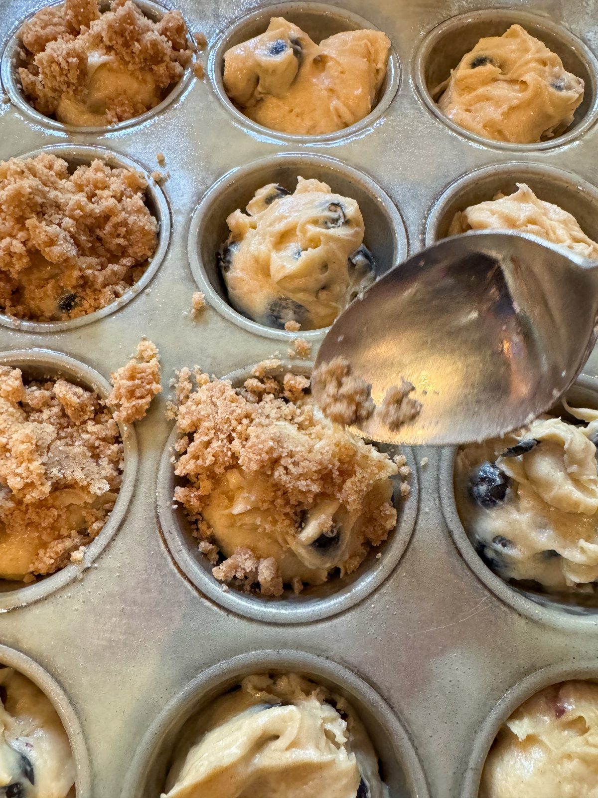 Recipe Photo - Blueberry Muffins with Cinnamon Streusel and Maple Glaze. Portrait orientation showing 12-cup muffin baking tin filled with scoops of batter and streusel topping being added with a spoon.