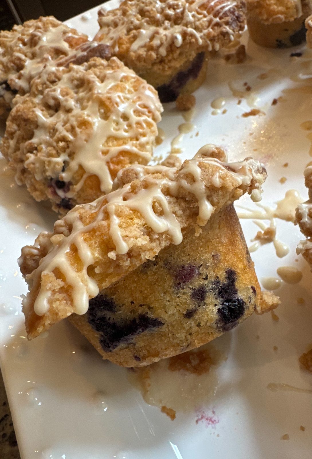 Recipe Photo - Blueberry Muffins with Cinnamon Streusel and Maple Glaze. Portrait orientation showing blueberry muffins on a platter. One muffin on its side showing the generous overflowing muffin top. Crumbs of streusel are scattered on the white platter.