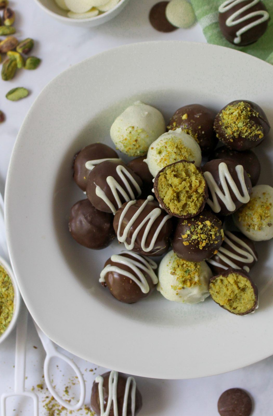Recipe beauty shot for Dubai Oreo Truffles. A white shallow bowl filled with dark, milk and white chocolate covered truffles. Some drizzled with chocolate and some sprinkled with pistachio dust. One is opened showing the greenish pistachio oreo filling.