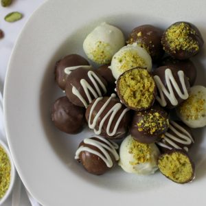 Recipe beauty shot for Dubai Oreo Truffles. A white shallow bowl filled with dark, milk and white chocolate covered truffles. Some drizzled with chocolate and some sprinkled with pistachio dust. One is opened showing the greenish pistachio oreo filling.