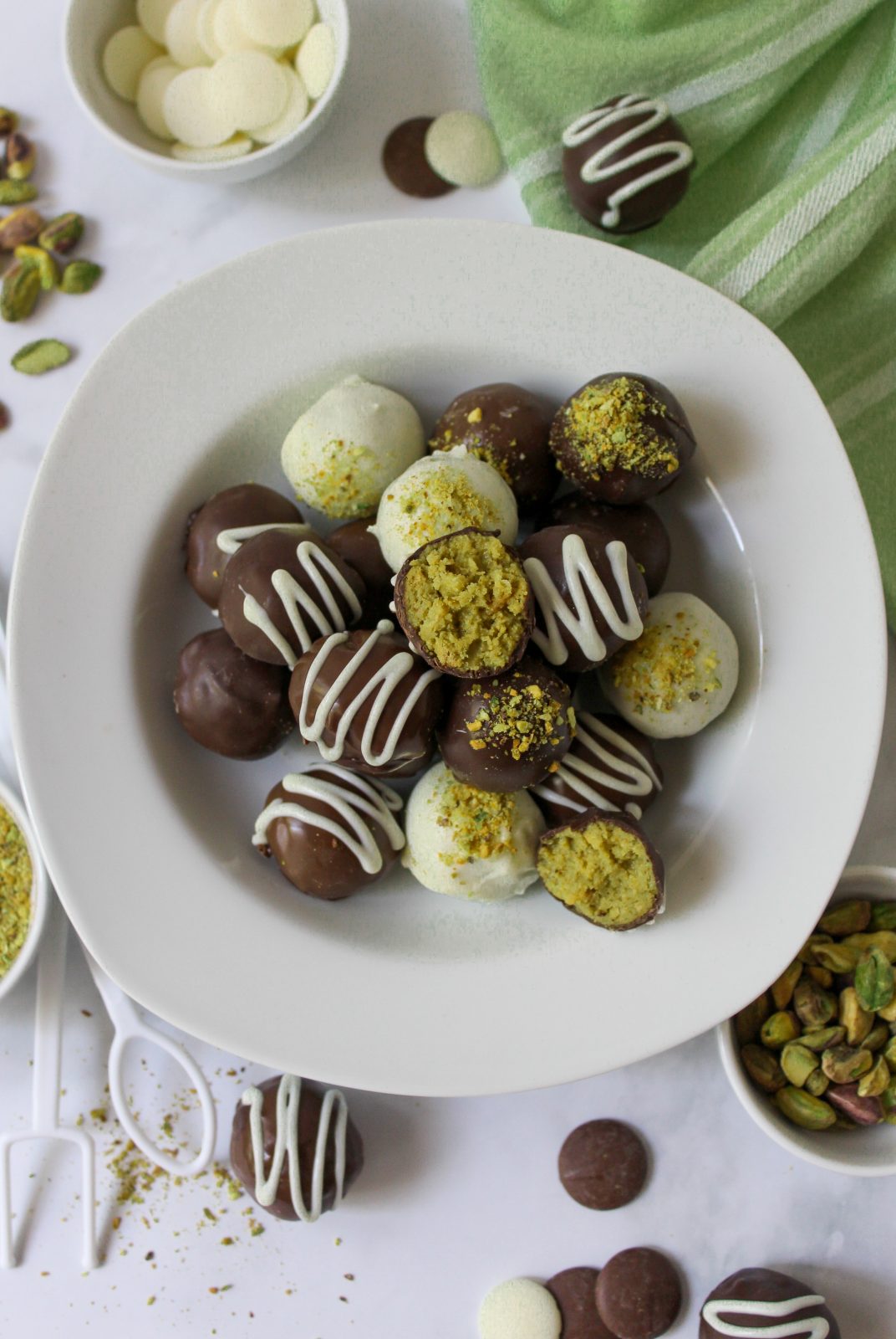 Recipe Beauty Shot of Dubai Style Pistachio Oreo Truffles. Bowl of dark, white and milk chocolate truffles decorated with pistachio dust or white chocolate drizzles in white shallow bowl. In the back ground are bowls of pistachios and chocolate. A few spilled pistachios. White plastic chocolate dipping tools and a green cloth napkin.