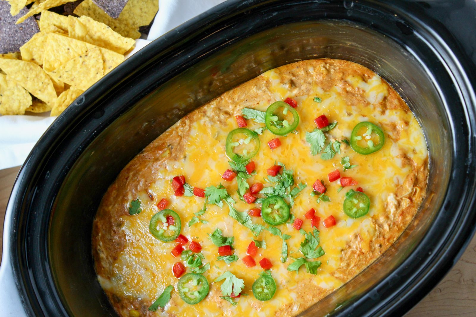 Recipe Photo for Chicken Enchilada Dip. Overhead shot of dip cooked in black oval stoneware slow cooker surrounded by multi colored tortilla chips. Dip is covered in cheddar cheese and sprinkled with sliced jalapeños, diced red bell peppers and a sprinkle of cilantro.