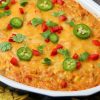 Recipe Photo for Chicken Enchilada Dip. Overhead shot of baked dip in oval stoneware baker surrounded by multi colored tortilla chips. Dip is covered in cheddar cheese and sprinkled with sliced jalapeños, diced red bell peppers and a sprinkle of cilantro.