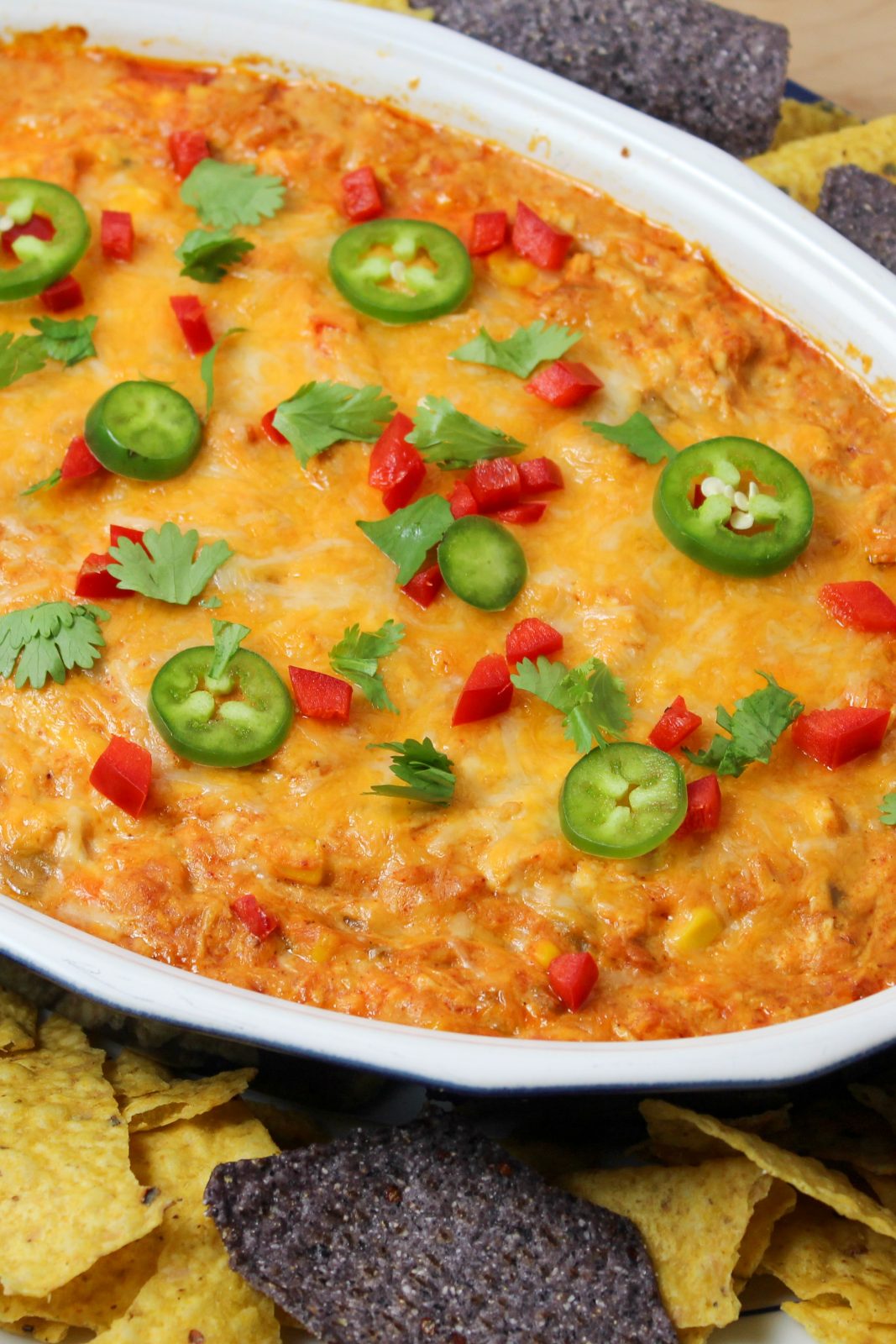 Recipe Photo for Chicken Enchilada Dip. Overhead shot of baked dip in oval stoneware baker surrounded by multi colored tortilla chips. Dip is covered in cheddar cheese and sprinkled with sliced jalapeños, diced red bell peppers and a sprinkle of cilantro.