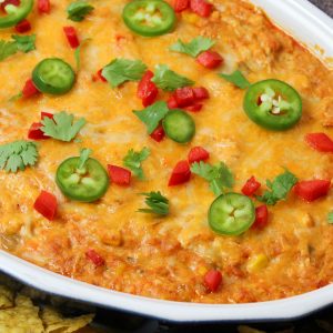 Recipe Photo for Chicken Enchilada Dip. Overhead shot of baked dip in oval stoneware baker surrounded by multi colored tortilla chips. Dip is covered in cheddar cheese and sprinkled with sliced jalapeños, diced red bell peppers and a sprinkle of cilantro.