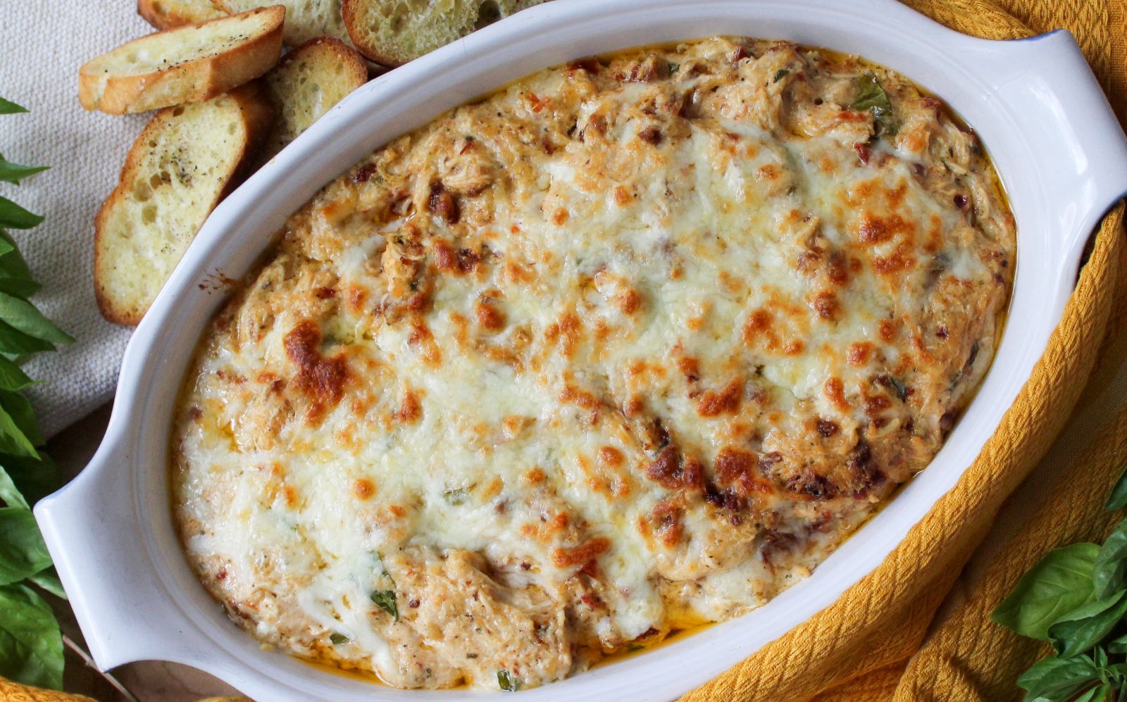 Recipe photo (landscape orientation) for Marry Me Chicken Dip. Showing final baked dip in an oval white stoneware baker, surrounded by a gold kitchen towel, fresh basil sprigs and toasted crostini. The dip is cheese topped and baked golden brown.