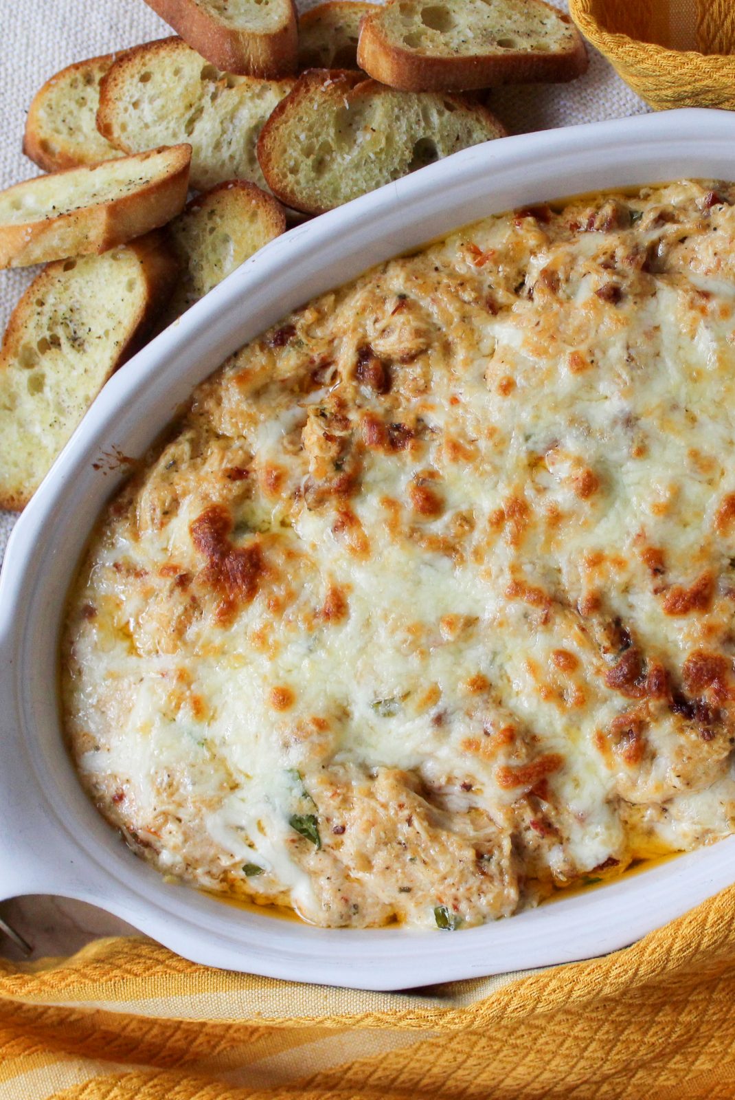 Recipe photo (portrait orientation) for Marry Me Chicken Dip. Showing final baked dip in an oval white stoneware baker, surrounded by a gold kitchen towel, fresh basil sprigs and toasted crostini. The dip is cheese topped and baked golden brown.