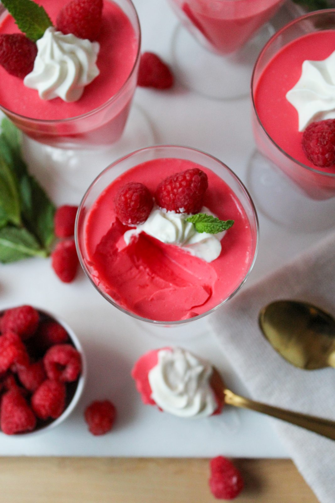 Recipe Photo for Creamy Greek Yogurt Jello.  Portrait orientation photo shows overhead shot of  4 tall footed parfait glasses filled with raspberry pink colored creamy opaque jello mixture, Topped with a swirl of piped whipped cream 2 raspberries and a fresh mint leaf.  The glasses are surrounded by some loose raspberries, mint sprigs and gold spoons.  There is a spoonful removed from the front glass.  The spoon is on the white marble the glasses sit atop.
