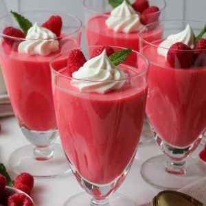 Recipe Photo for Creamy Greek Yogurt Jello. Portrait orientation photo shows 4 tall footed parfait glasses filled with raspberry pink colored creamy opaque jello mixture, Topped with a swirl of piped whipped cream 2 raspberries and a fresh mint leaf. The glasses are surrounded by some loose raspberries and gold spoons