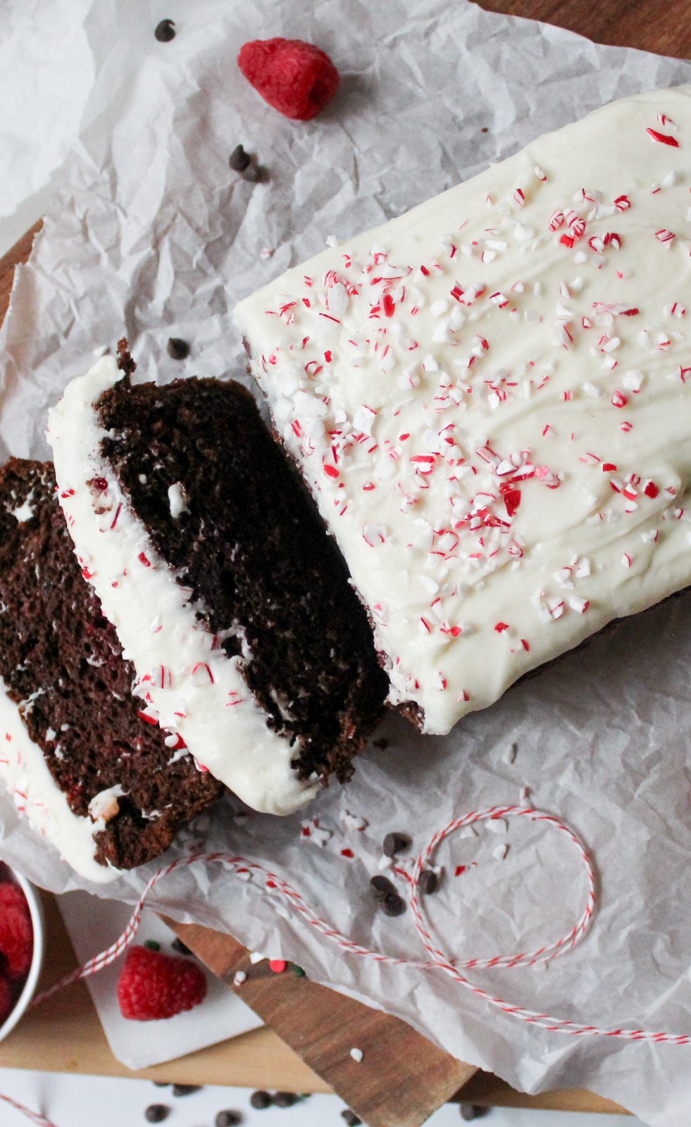Recipe Photo for Easy Chocolate Loaf Cake. Overhead close up portrait shot of chocolate loaf cake on parchment paper atop a wooden cutting board. 2 slices cut showing moist interior. Top is covered in cream cheese glaze and peppermint sprinkles for the peppermint bark version of the recipe. There are sprinkles. chocolate chips, baking twine and raspberries scattered around the cake.