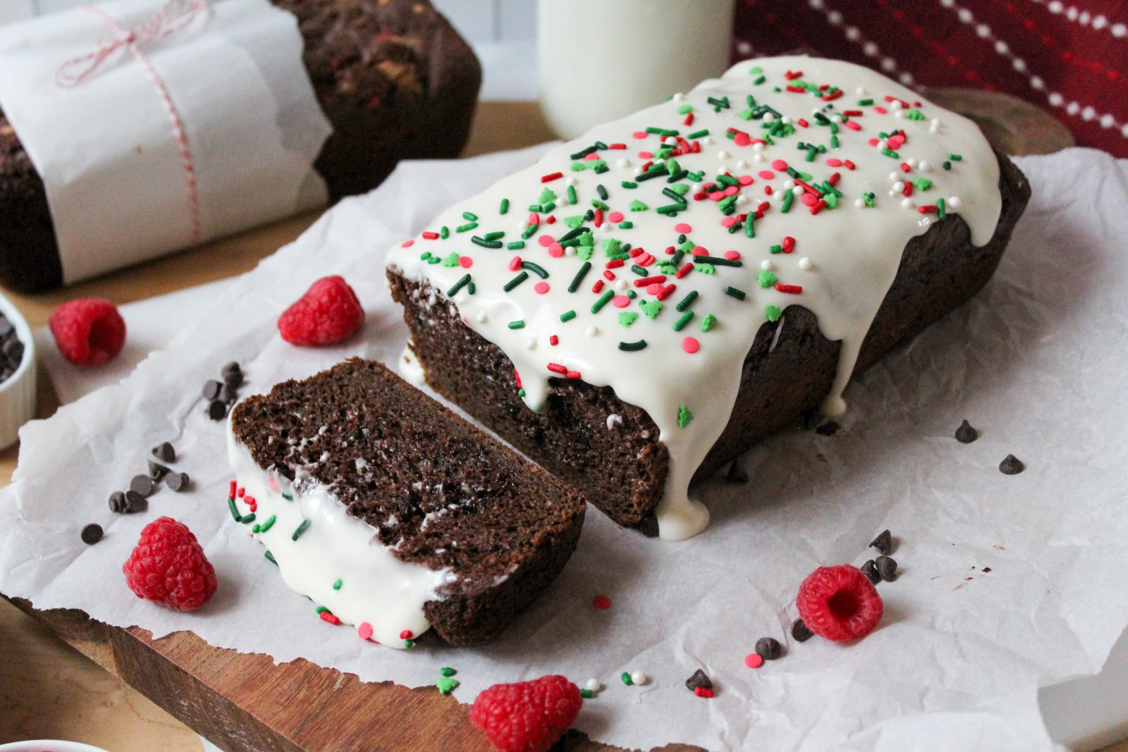 Recipe Photo for Easy Chocolate Loaf Cake. 45 degree angle landscape shot of chocolate loaf cake on parchment paper atop a wooden cutting board. 2 slices cut showing moist interior. Top is covered in cream cheese glaze and festive sprinkles. There are sprinkles. chocolate chips, baking twine and raspberries scattered around the cake.