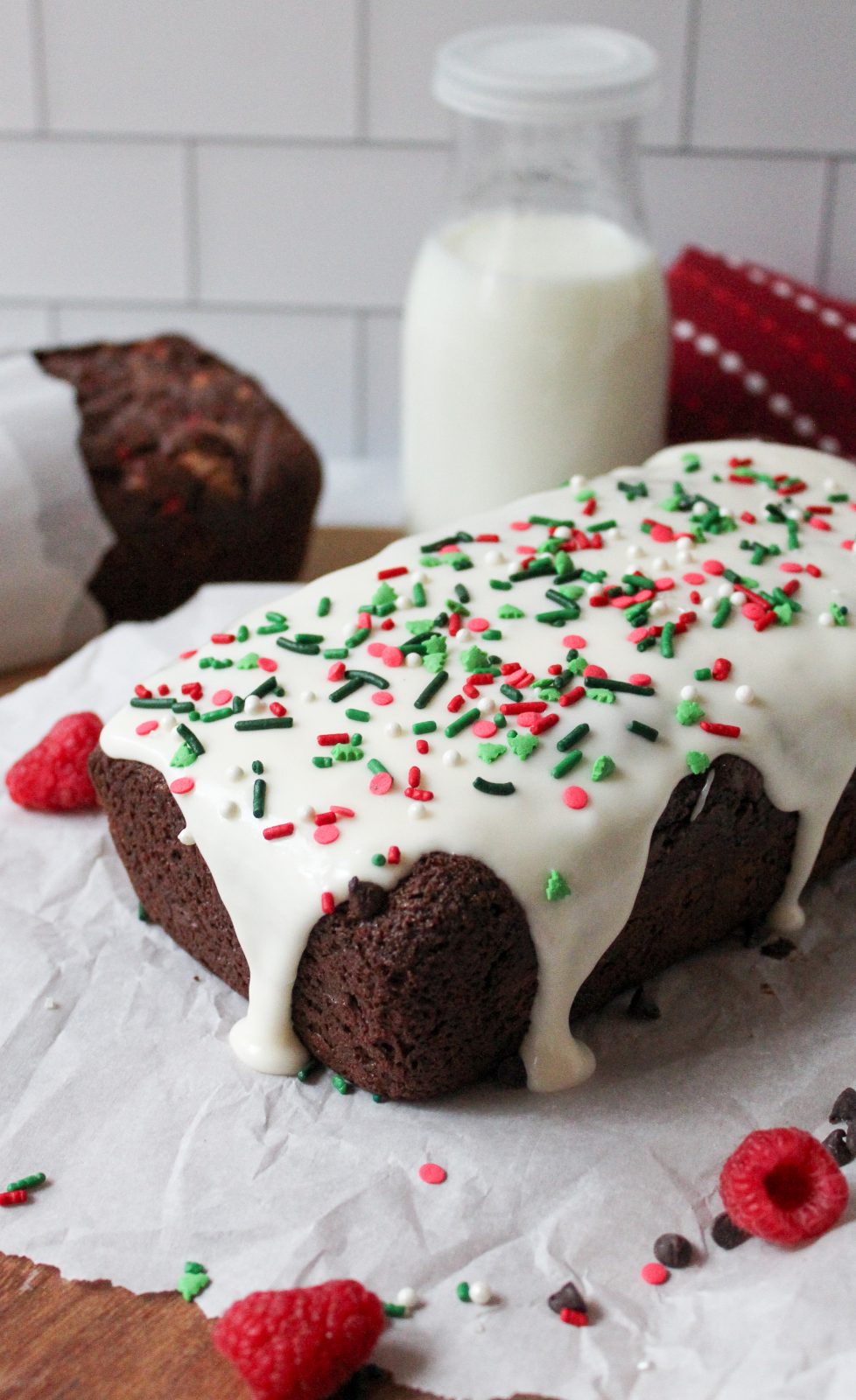 Recipe Photo for Easy Chocolate Loaf Cake. 45 degree angle landscape shot of chocolate loaf cake on parchment paper atop a wooden cutting board. Top is covered in cream cheese glaze and festive sprinkles. There are sprinkles. chocolate chips, baking twine and raspberries scattered around the cake.