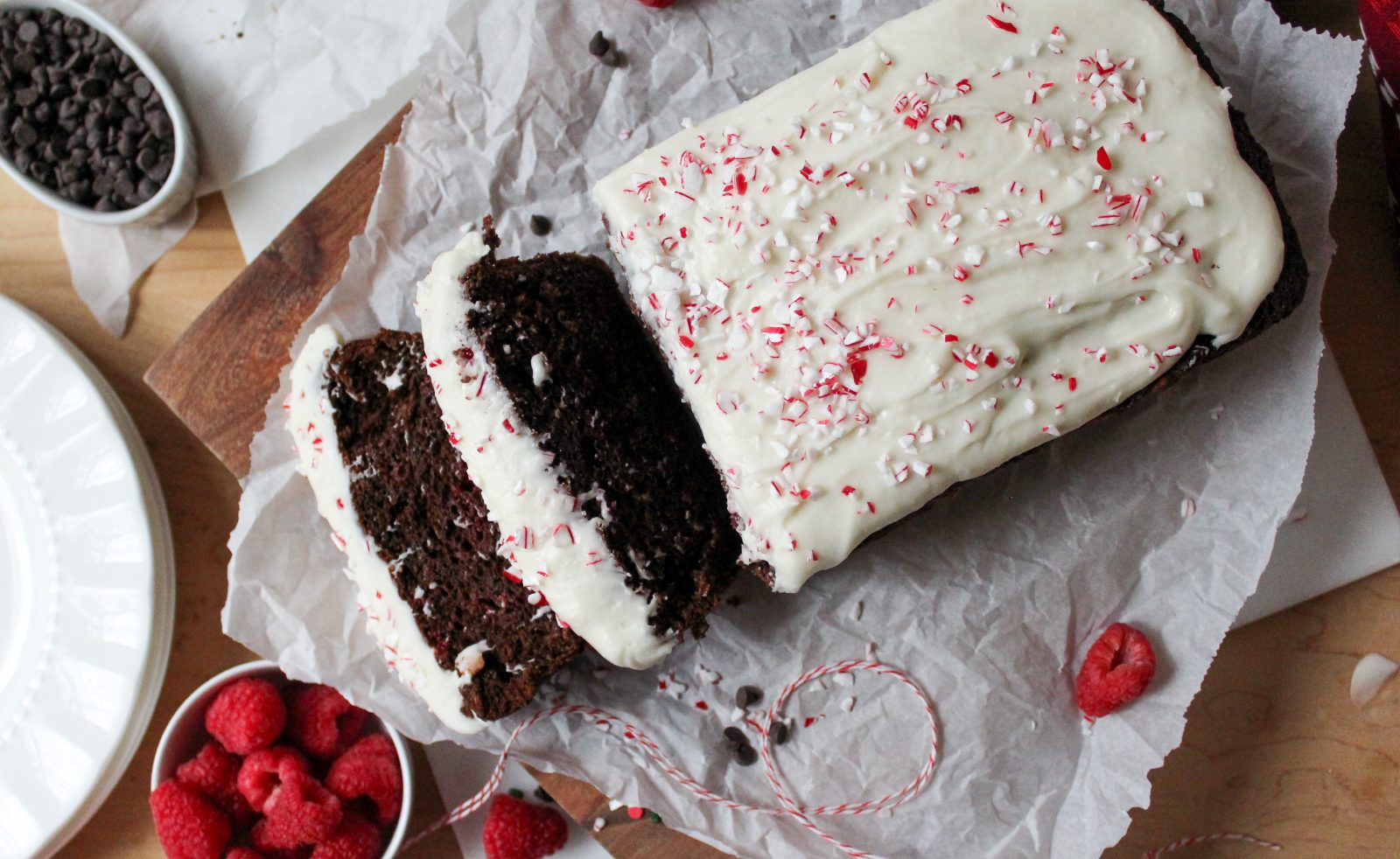 Recipe Photo for Easy Chocolate Loaf Cake. Overhead landscape shot of chocolate loaf cake on parchment paper atop a wooden cutting board. 2 slices cut showing moist interior. Top is covered in cream cheese glaze and peppermint sprinkles for the peppermint bark version of the recipe. There are sprinkles. chocolate chips, baking twine and raspberries scattered around the cake along with some plates.