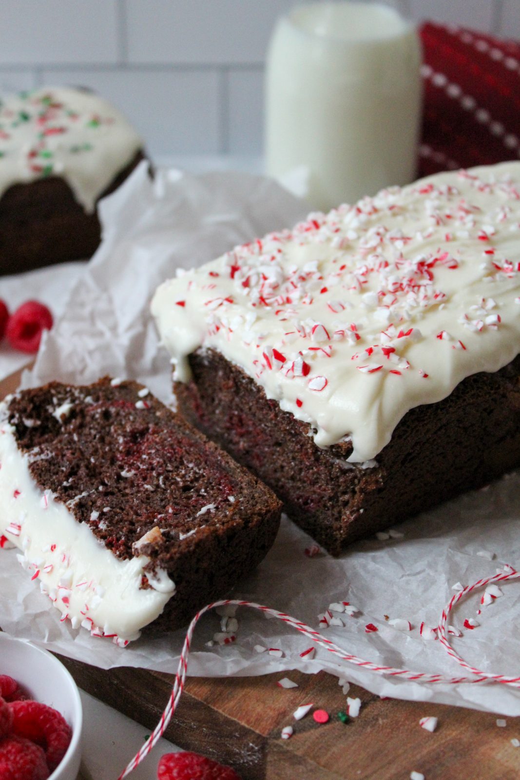 Recipe Photo for Easy Chocolate Loaf Cake. 45 degree angle portrait shot of chocolate loaf cake on parchment paper atop a wooden cutting board. 2 slices cut showing moist interior. Top is covered in cream cheese glaze and peppermint sprinkles for the peppermint bark version of the recipe. There are sprinkles. chocolate chips, baking twine and raspberries scattered around the cake.
