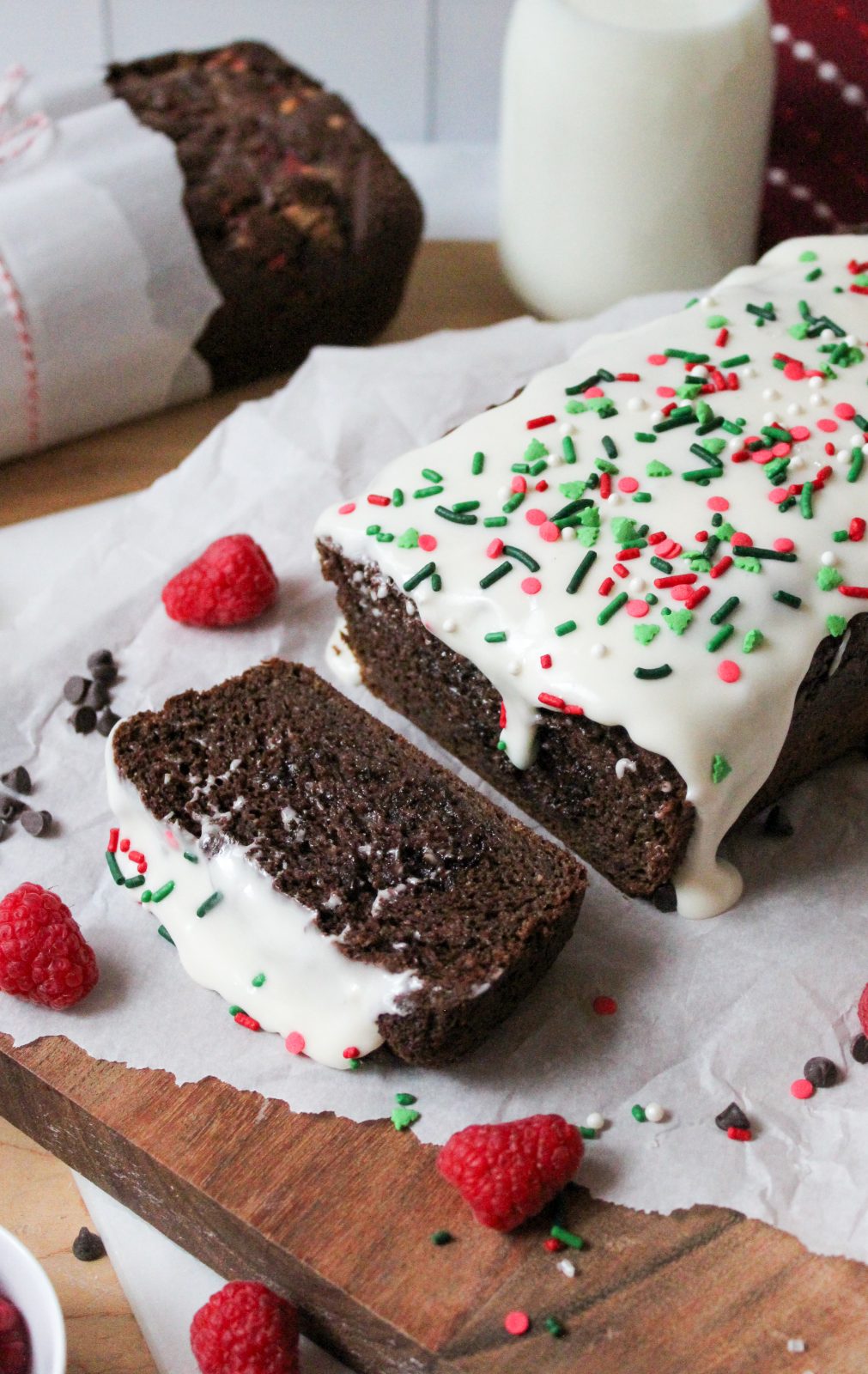 Recipe Photo for Easy Chocolate Loaf Cake. 45 degree angle portrait shot of chocolate loaf cake on parchment paper atop a wooden cutting board. 2 slices cut showing moist interior. Top is covered in cream cheese glaze and festive sprinkles. There are sprinkles. chocolate chips, baking twine and raspberries scattered around the cake.