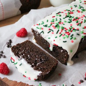 Recipe Photo for Easy Chocolate Loaf Cake. 45 degree angle portrait shot of chocolate loaf cake on parchment paper atop a wooden cutting board. 2 slices cut showing moist interior. Top is covered in cream cheese glaze and festive sprinkles. There are sprinkles. chocolate chips, baking twine and raspberries scattered around the cake.