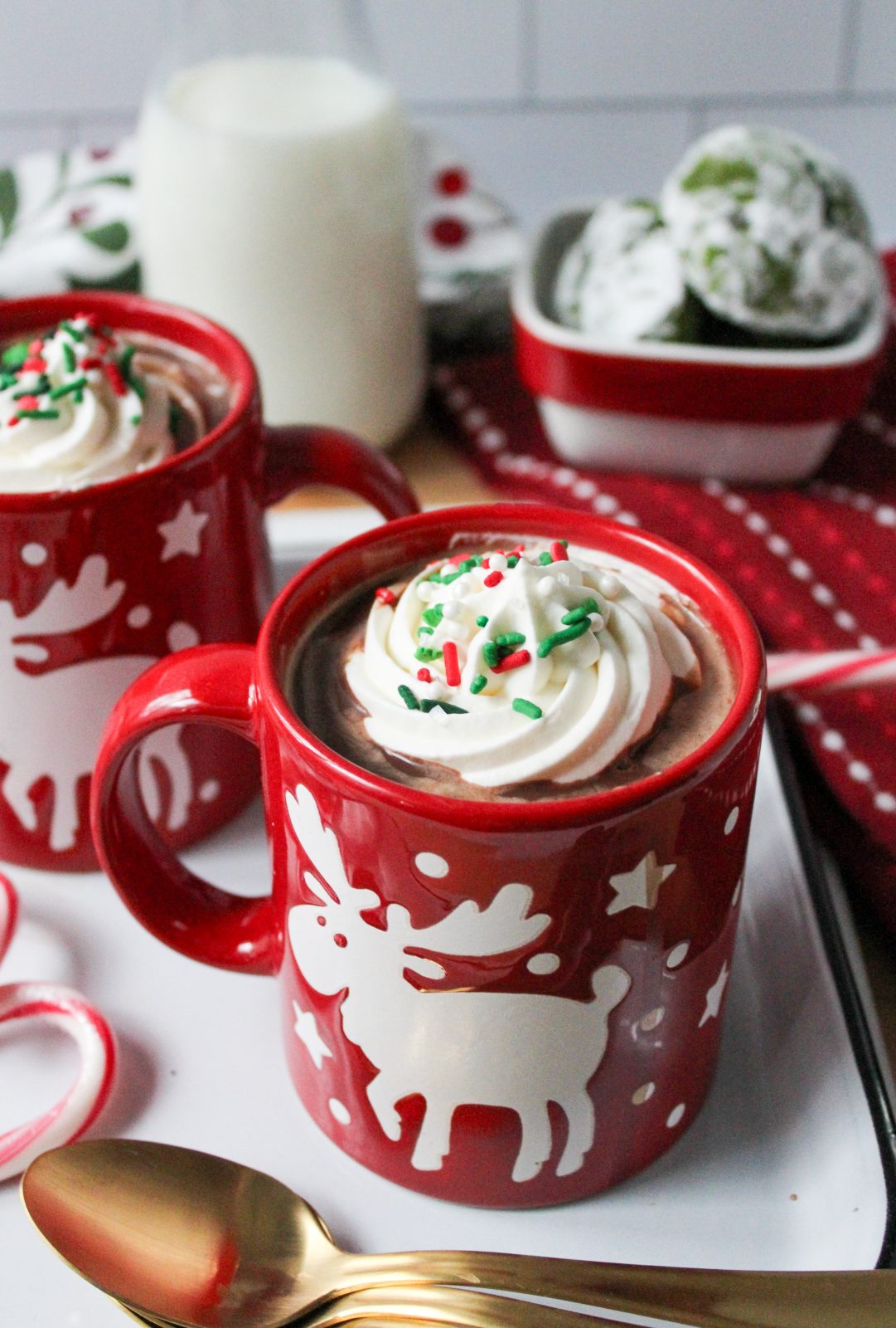 Recipe Photo for Frozen Marshmallow Whipped Cream Cocoa Toppers. Portrait orientation photo with wide angle shows red moose mugs with cocoa and the frozen toppers floating on the cocoa. The mugs are on a vintage enamel tray with gold spoons and candy canes. In the background is a small bowl with green crinkle cookies and a small jar with milk. You can see a white subway backsplash in the background.
