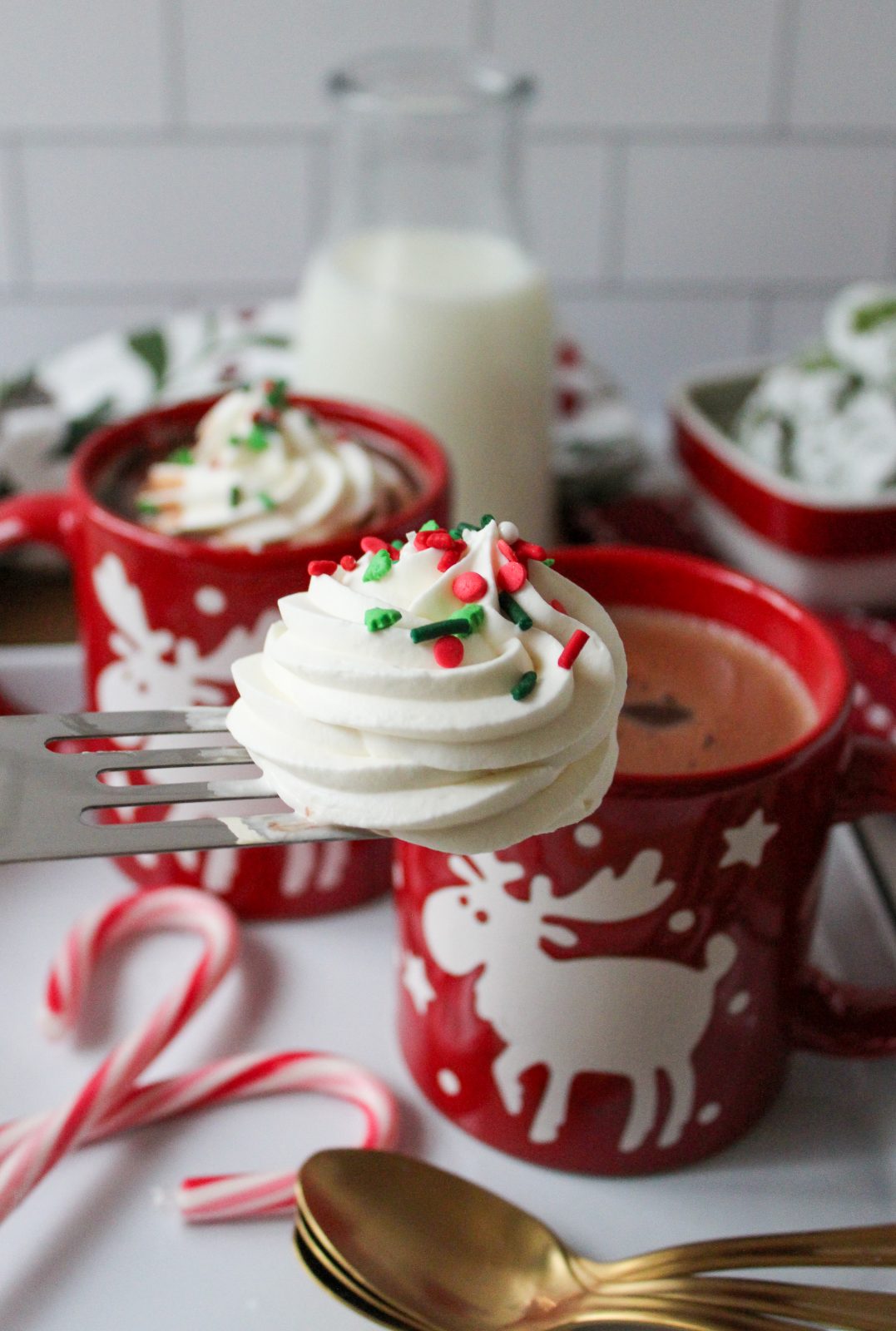 Recipe Photo for Frozen Marshmallow Whipped Cream Cocoa Toppers. Portrait orientation photo shows red moose mugs with cocoa and a spatula transferring the frozen topper onto the cocoa. The mugs are on a vintage enamel tray with gold spoons and candy canes. In the background is a small bowl with green crinkle cookies and a small jar with milk.