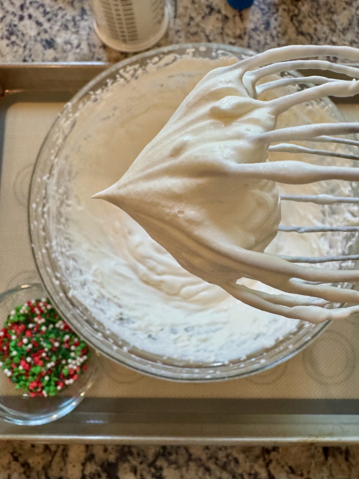 Recipe Photo for Frozen Marshmallow Whipped Cream Cocoa Toppers. Portrait orientation photo shows a close up of a whisk with a stiff peak of whipped cream overtop a glass bowl of the mixture.