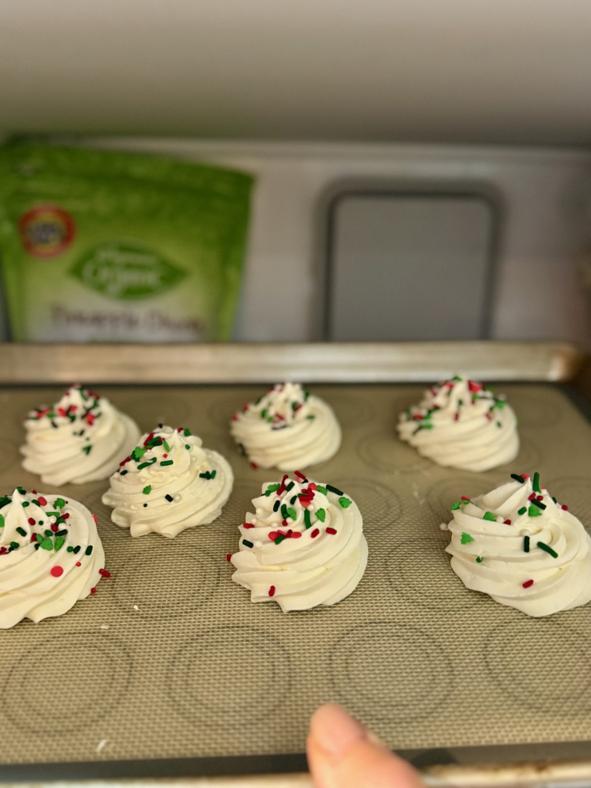 Recipe Photo for Frozen Marshmallow Whipped Cream Cocoa Toppers. Portrait orientation photo shows a silicone mat with 7 piped rounds made using a star piping tip of the whipped cream mixture. The rounds on the tray are being taken out of a freezer. There is a frozen bag of something in the background of the freezer.
