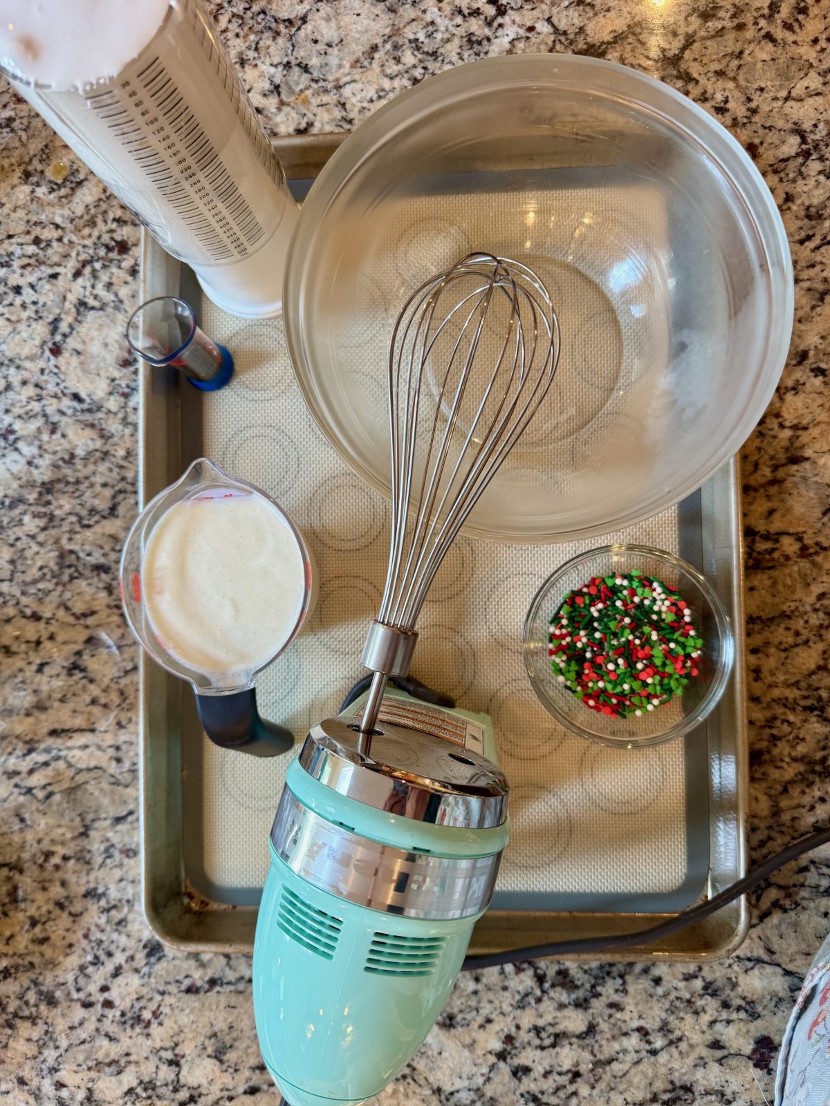 Recipe Photo for Frozen Marshmallow Whipped Cream Cocoa Toppers. Portrait orientation photo shows Ingredients for making recipe on a silicone mat lined sheet tray. Heavy Cream, marshmallow fluff, vanilla and sprinkles. There is also an Ice Blue KitchenAid Hand Mixer in the picture with a Whisk beater attached.