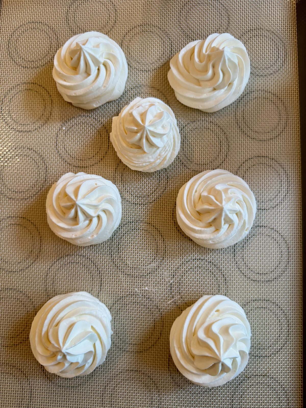 Recipe Photo for Frozen Marshmallow Whipped Cream Cocoa Toppers. Portrait orientation photo shows a silicone mat with 7 piped rounds using a star piping tip of the whipped cream mixture.