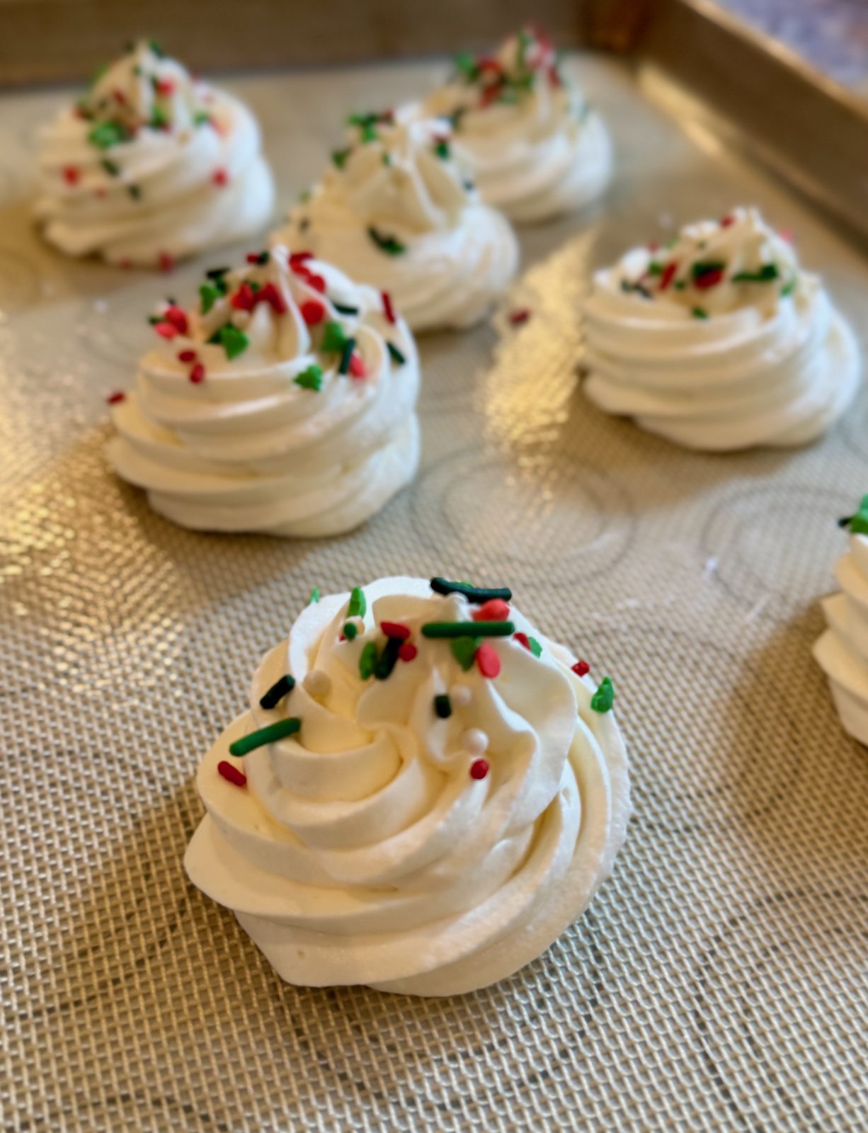 Recipe Photo for Frozen Marshmallow Whipped Cream Cocoa Toppers. Portrait orientation photo shows a silicone mat with 6 piped rounds using a star piping tip of the whipped cream mixture. The toppers are sprinkled with Holiday red and green various shaped sprinkles.