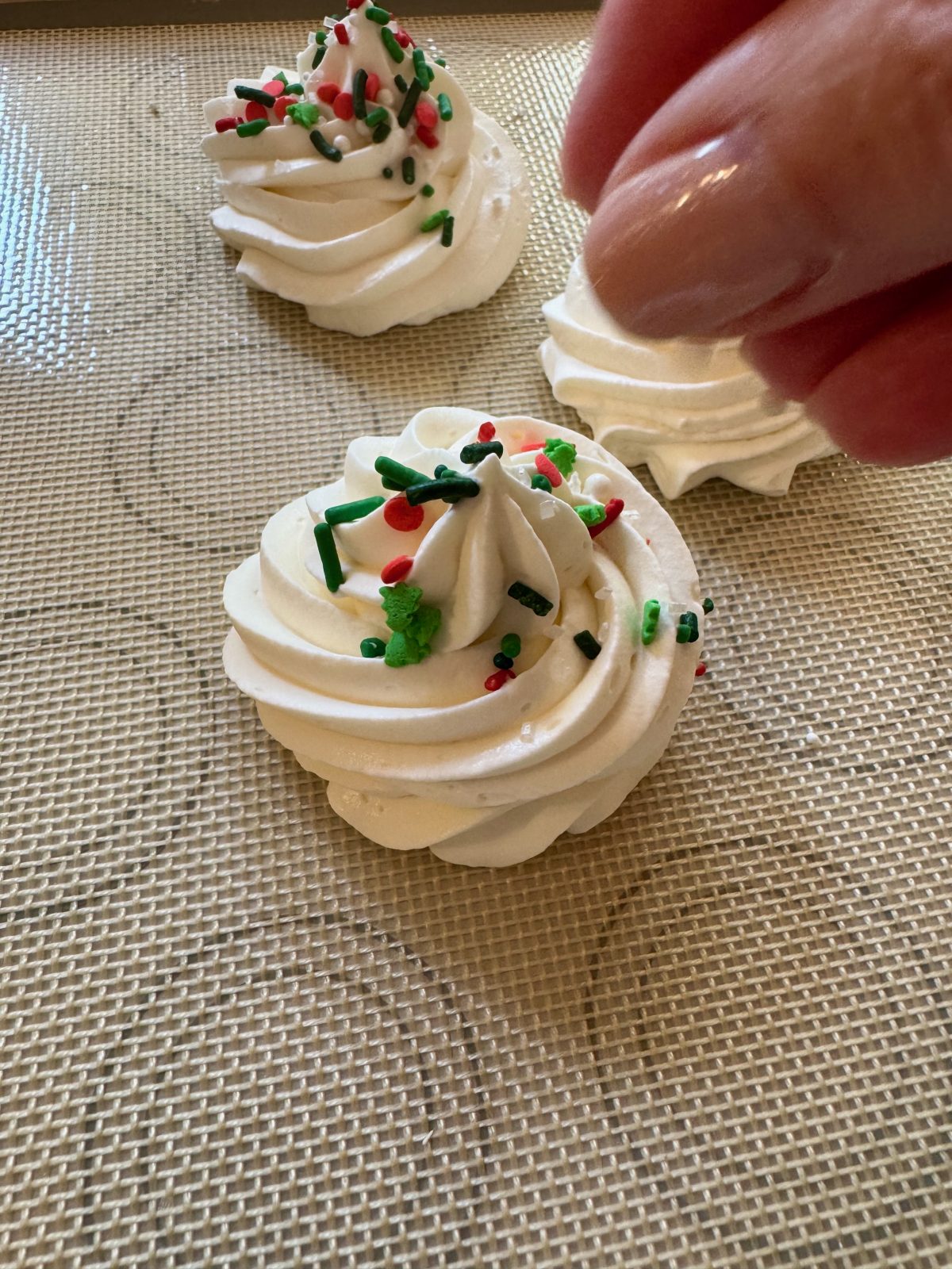 Recipe Photo for Frozen Marshmallow Whipped Cream Cocoa Toppers. Portrait orientation photo shows a silicone mat with 3 piped rounds using a star piping tip of the whipped cream mixture. A hand is sprinkling Holiday red and green various shaped sprinkles on one of the toppers.