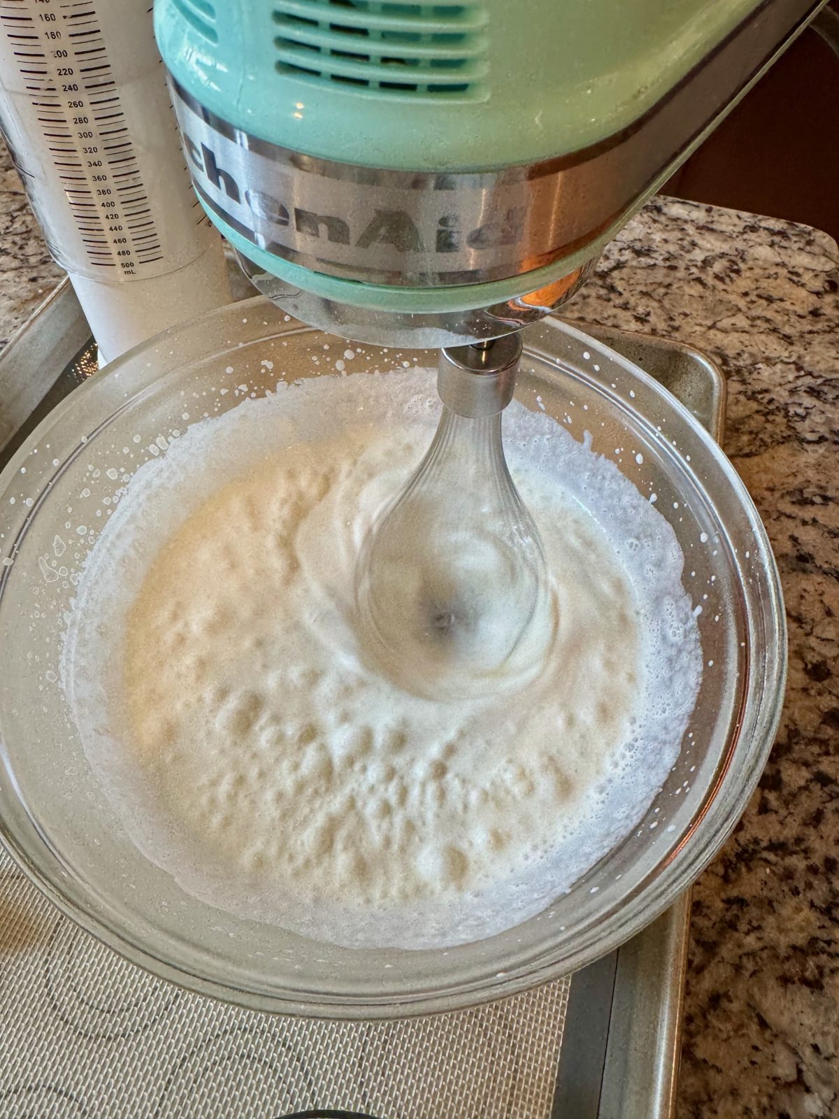 Recipe Photo for Frozen Marshmallow Whipped Cream Cocoa Toppers. Portrait orientation photo shows an Ice Blue KitchenAid Hand Mixer in with a Whisk beater attached whipping cream in a chilled glass bowl.