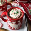 Recipe Photo for Frozen Marshmallow Whipped Cream Cocoa Toppers. Portrait orientation photo shows red moose mugs with cocoa and the frozen toppers floating on the cocoa. The mugs are on a vintage enamel tray with gold spoons and candy canes. In the background is a small bowl with green crinkle cookies and a small jar with milk.